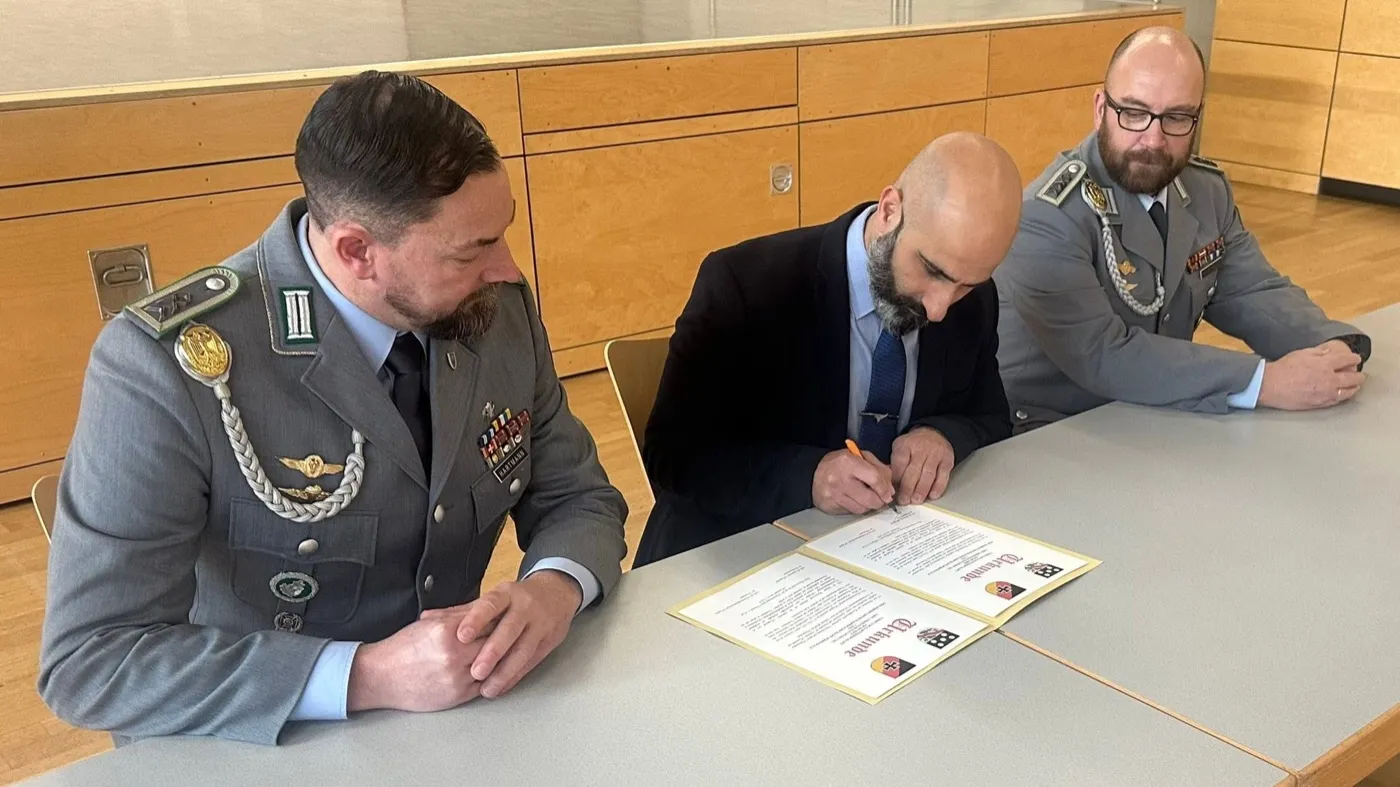 Drei Männer an einem Tisch in einem hellen Saal: Nader Samadi Tehrani in dunklem Anzug unterschreibt eine aufgeschlagene Urkunde, links und rechts von ihm je ein Soldat im Dienstanzug der Bundeswehr