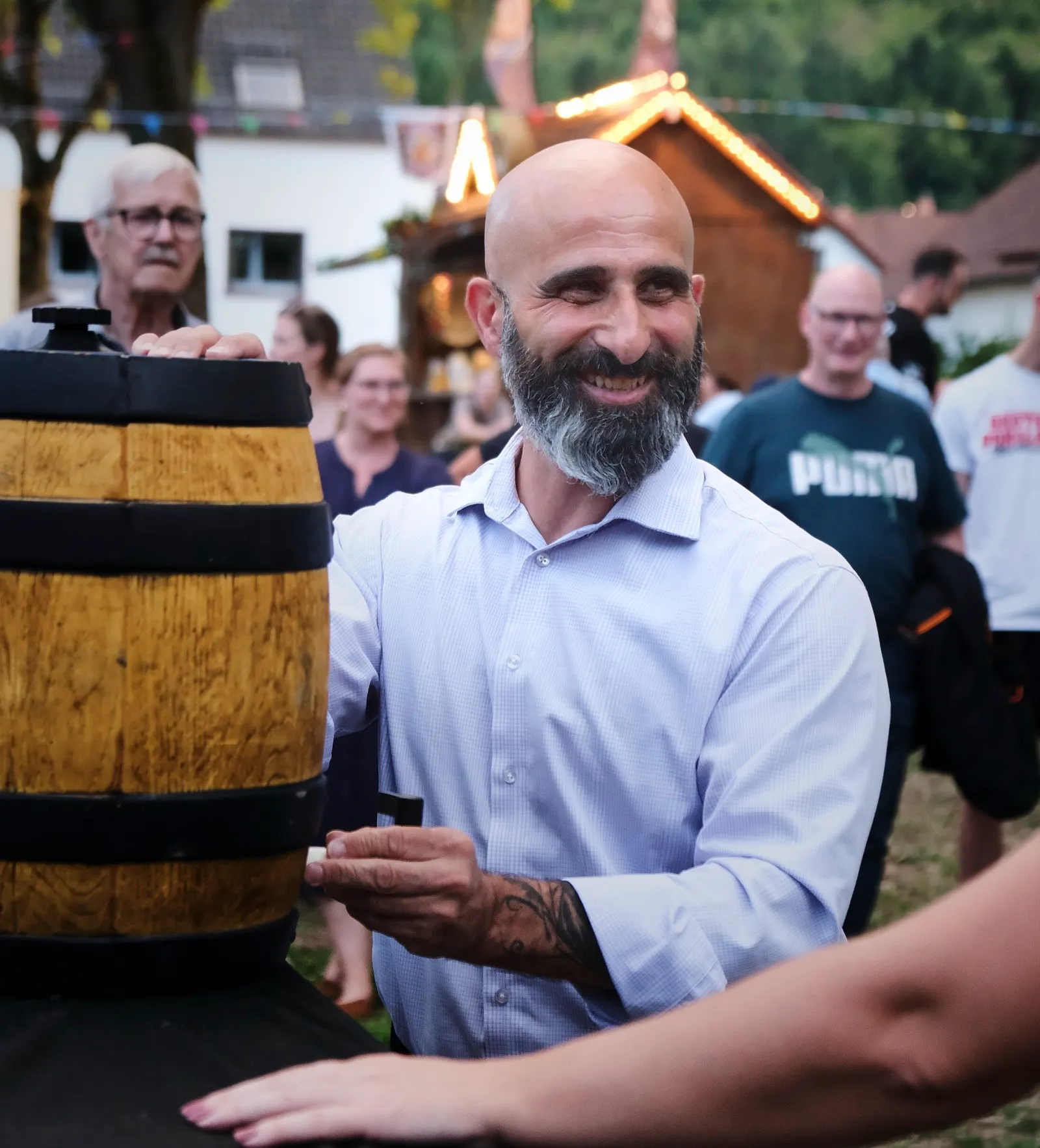 Nader Samadi Tehrani im hellblauen Hemd an einem hölzernen Fass. Eine Hand liegt auf dem Fassdeckel, in der anderen hält er den Zapfhahn. Er lächelt und blickt auf das Fass. Im Hintergrund stehen Gäste bei einer geschmückten Hütte und Lichterketten.